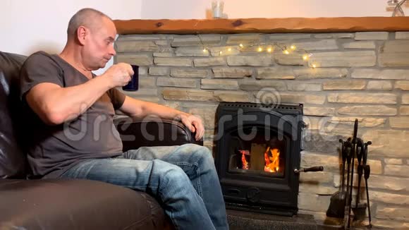 一个喝着咖啡和茶的中年男人坐在一块烧着的石头旁边的皮革沙发上享受着温暖有感情地微笑着视频的预览图