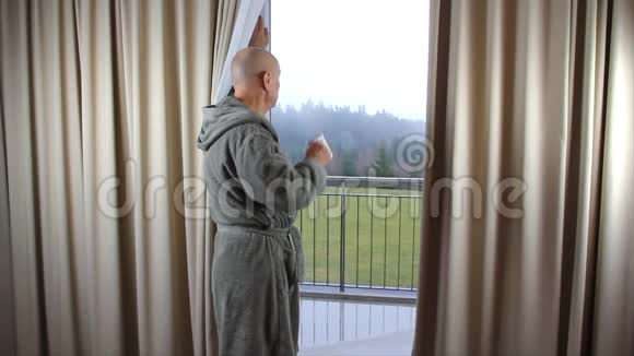 穿着灰色浴袍的中年男子从窗户边的杯子里喝茶或咖啡视频的预览图