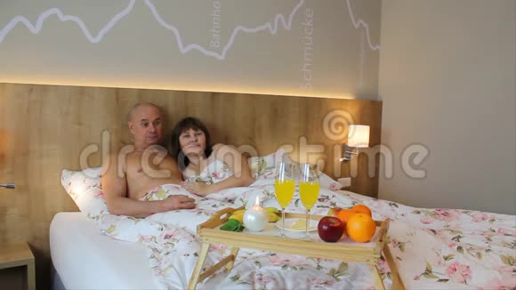 微笑的女人和男人躺在酒店房间的床上在一张桌子前集中着水果和饮料早餐在床上视频的预览图