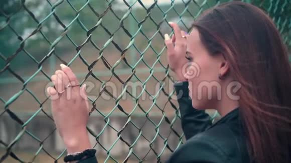 漂亮而热情的女孩穿着铁栅栏后面的自行车夹克被关在监狱里的年轻女子锁着金属栅栏视频的预览图
