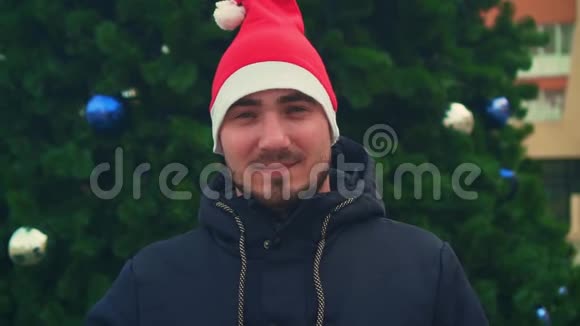 有胡子的年轻人戴着圣诞帽背景是圣诞树快乐的男人用超级手展示标志视频的预览图