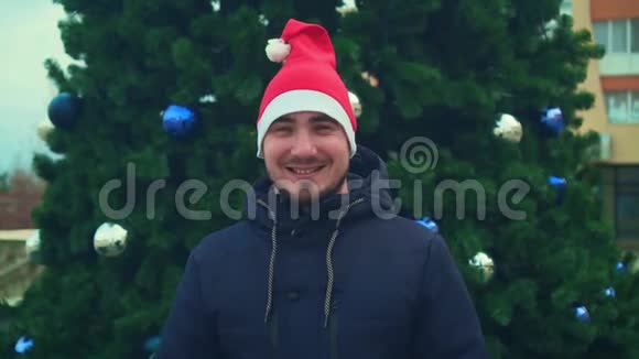 有胡子的年轻人戴着圣诞帽背景是圣诞树快乐的男人对着镜头微笑视频的预览图