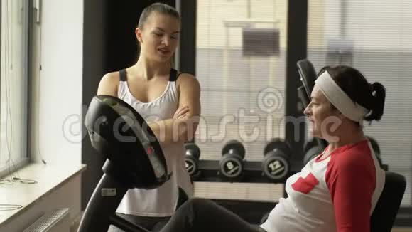 中年妇女在健身房骑自行车模拟器锻炼而女教练则站在他旁边视频的预览图
