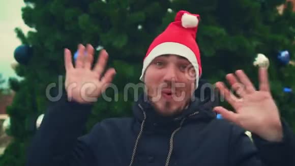 有胡子的年轻人戴着圣诞帽背景是圣诞树一个快乐的人挥舞着双手跳舞露出他的舌头视频的预览图
