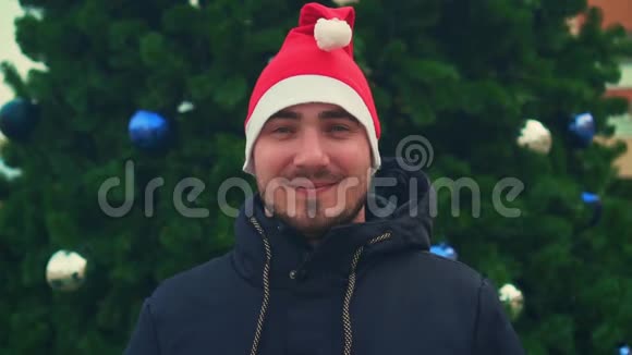 有胡子的年轻人戴着圣诞帽背景是圣诞树一个快乐的人用两只手展示一个超级标志视频的预览图