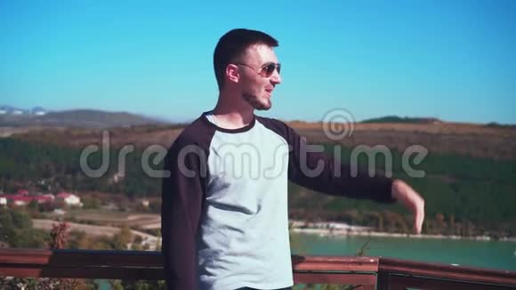 一个戴眼镜的年轻性感男子站在观景台上俯瞰湖面那人向远处微笑举起手来视频的预览图