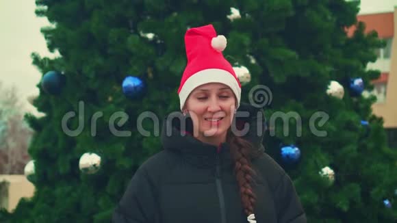 圣诞树背景上戴圣诞帽的年轻女子快乐的漂亮女人微笑着挥舞着手跳舞视频的预览图