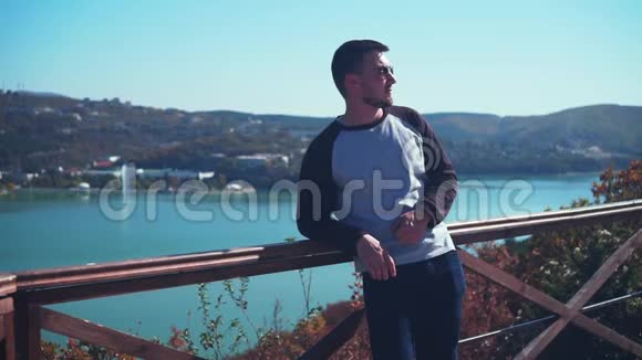 戴眼镜的年轻性感男人在俯瞰湖面的观景台上一个男人为摄像机摆姿势视频的预览图