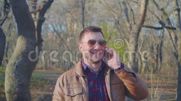 穿着皮夹克棒球帽和时髦太阳镜在智能手机上说话的人的肖像他微笑着视频的预览图