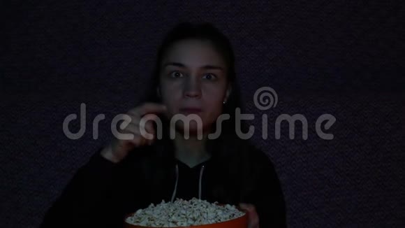 一个看电视吃爆米花的年轻女孩年轻漂亮的女孩坐在家里的沙发上看她最喜欢的电视电影视频的预览图