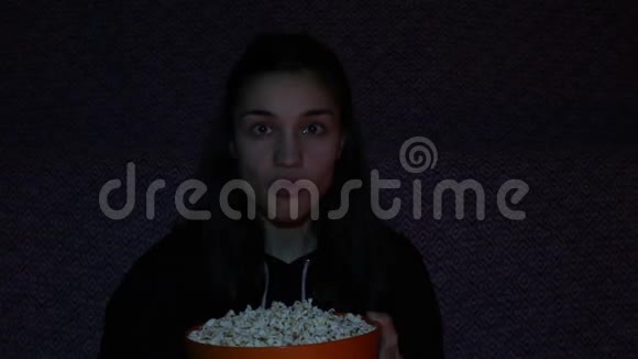 一个看电视吃爆米花的年轻女孩年轻漂亮的女孩坐在家里的沙发上看她最喜欢的电视电影视频的预览图