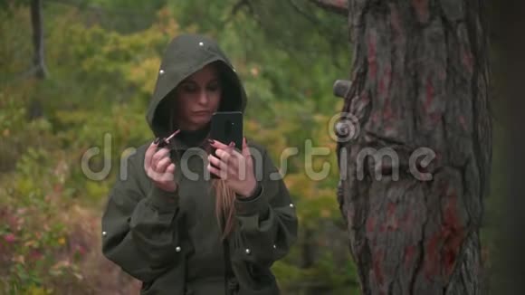 穿灰色夹克的漂亮女孩在森林背景上看智能手机视频的预览图
