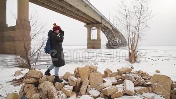 一个女孩在桥附近拍摄一条冰冻的河流冬天女游客在大自然中拍照视频的预览图