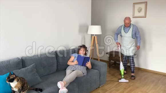 55岁的秃头男人穿着家居服在沙发附近的房间里用围裙吸尘地板女人男人坐在沙发上而不是视频的预览图