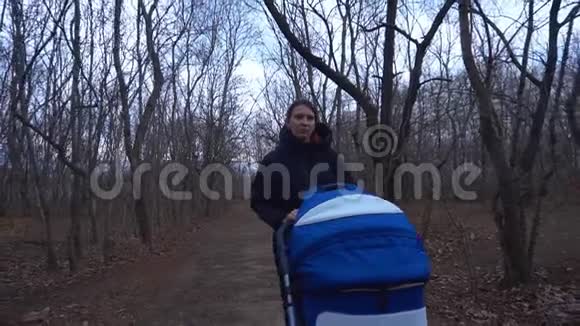 一个女人带着婴儿车在公园里散步一个穿着温暖夹克和帽子的女人在树林里卷着婴儿车这就是视频的预览图