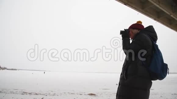 女游客在桥下的河边拍摄一幅冬天的风景女摄影师在大自然中拍照视频的预览图