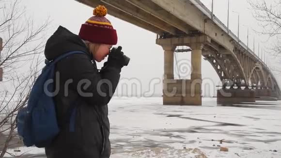 女游客在桥下的河边拍摄一幅冬天的风景女摄影师在大自然中拍照视频的预览图