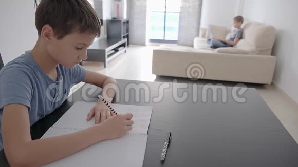 专注的男孩坐在桌子旁写数学作业他的哥哥坐在沙发上用笔记本电脑视频的预览图