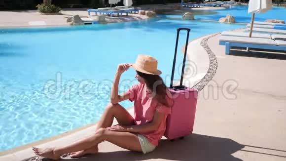 带粉红色箱子的年轻女子坐在酒店游泳池附近旅游度假概念高清视频支持1920x1080视频的预览图