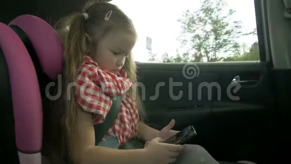 可爱的小女孩一边忙着用智能手机一边坐在车的后座上可爱的小学儿童玩耍视频的预览图