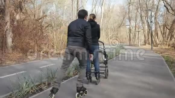 活泼开朗的爸爸和妈妈穿着溜冰鞋在公园的小巷里散步带着孩子在婴儿车里后景视频的预览图