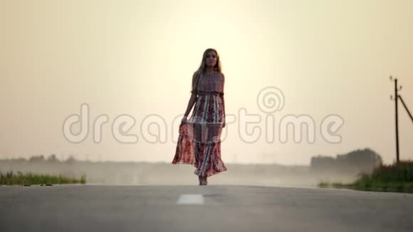 一个年轻漂亮的女孩穿着一件长裙子在一个夏天温暖的夜晚赤脚走在路上视频的预览图