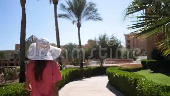 美丽的女人戴着白色的大帽子和粉红色的旅行袋走在酒店的领地上她用棕榈树在小巷里散步视频的预览图