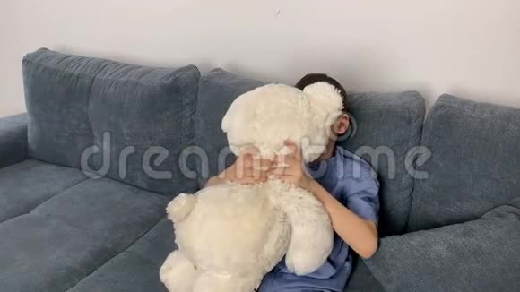 孩子一个小学年龄的男孩穿着牛仔裤一件蓝色衬衫坐在沙发上拥抱一只白色的泰迪熊孩子的概念s视频的预览图