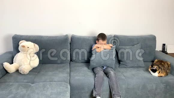 孩子一个穿着牛仔裤的小学小男孩一件蓝色衬衫坐在沙发上遮住了他的脸一只白色的泰迪在附近视频的预览图