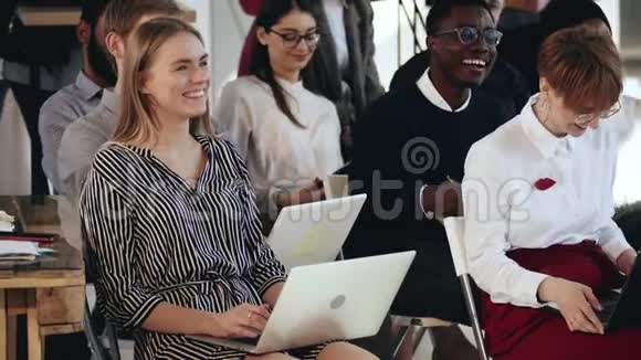 快乐的年轻金发商务女性坐着多民族的团队和笔记本电脑在现代办公室听着微笑的研讨会视频的预览图