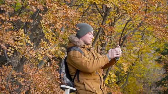 年轻的男游客戴着一顶帽子穿着一件带背包的夹克在秋天森林的背景上合影留念视频的预览图