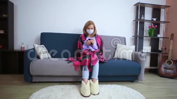 女孩病了坐在家里的沙发上戴着防护面具手里拿着一个杯子裹着一种格子病毒视频的预览图