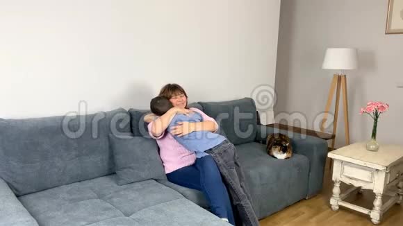 妈妈坐在沙发上孩子一个小学年龄的男孩穿着牛仔裤一件蓝色衬衫站在他旁边拥抱她育儿的概念视频的预览图