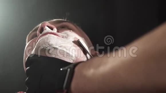 戴黑色手套的理发师非常小心地用危险的剃刀剃掉客户的胡须黑色背景视频的预览图