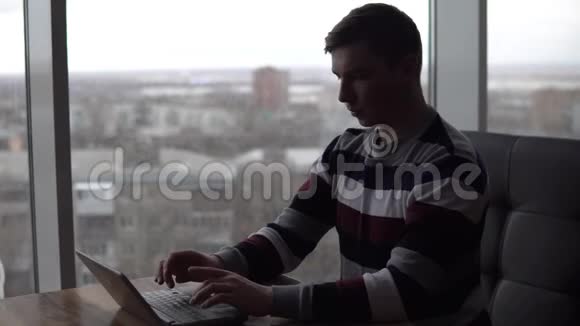 一个拿着笔记本电脑的年轻人坐在桌子旁一个男人坐在全景窗口旁的咖啡馆里手里拿着一台小笔记本电脑写着视频的预览图