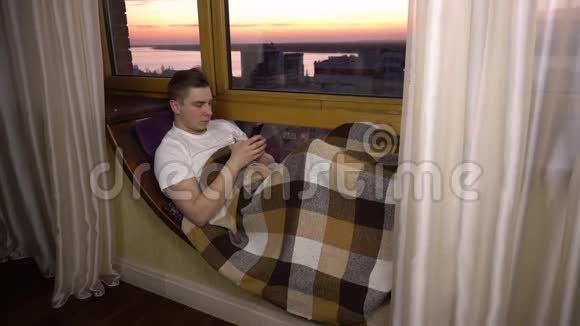 一个年轻人正在打电话一个男人躺在窗边的窗台上手里拿着一部智能手机外部视频的预览图
