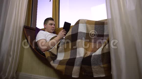 一个年轻人在平板电脑上发短信一个人躺在窗边的窗台上手里拿着一块平板电脑在外面视频的预览图