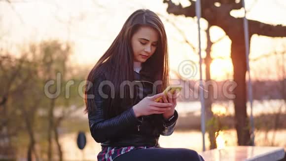 一位美丽的女性在镜头前的日落时分坐在公园里用她的手机集中着视频的预览图