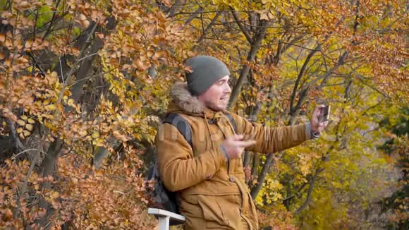 年轻的男游客戴着一顶帽子穿着一件背包的夹克挥舞着手通过智能手机上的视频交流交谈视频的预览图