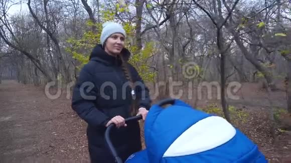 秋天的森林里一个女人带着婴儿车在公园里散步一个穿暖和夹克和帽子的女人在婴儿车上翻滚视频的预览图