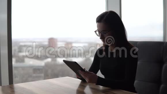 一个拿着平板电脑的年轻女人坐在桌子旁一个女孩坐在全景窗旁的咖啡馆里手里拿着一块平板电脑视频的预览图