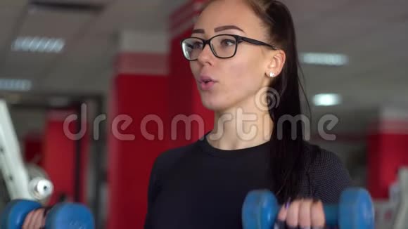 年轻的健身妇女在健身房用蓝色哑铃锻炼体育俱乐部戴眼镜的漂亮女孩视频的预览图