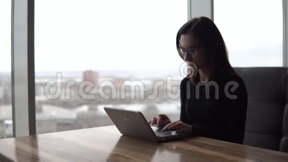 一个年轻的女人带着笔记本电脑坐在桌子旁一个女孩坐在全景窗口旁边的咖啡馆里手提电脑和视频的预览图