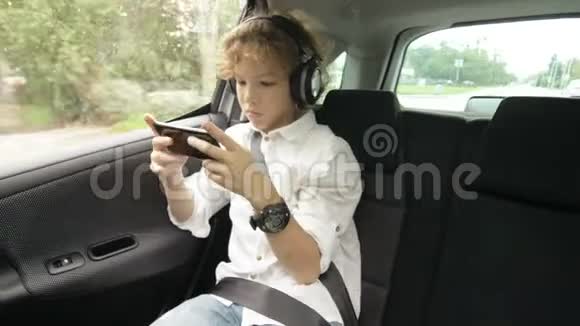 带着耳机的十几岁男孩坐在带安全带的车里用智能手机视频的预览图