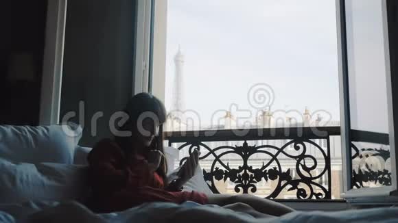 美丽的快乐博客女性躺在法国阳台窗口与埃菲尔铁塔景使用智能手机慢动作视频的预览图