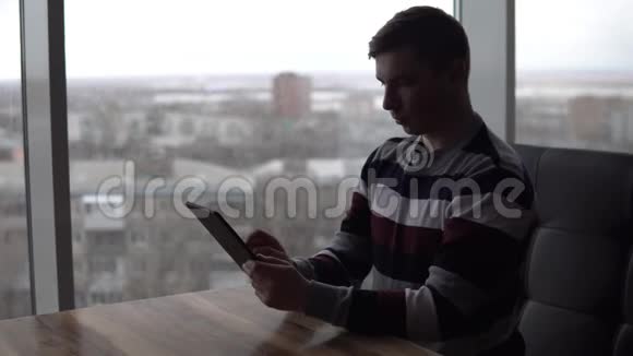 一个拿着平板电脑的年轻人坐在桌子旁一个男人坐在全景窗旁的咖啡馆里手里拿着一块平板电脑视图视频的预览图