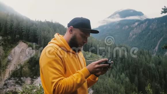 年轻的快乐胡子男子穿着黄色连帽衫正在控制无人机飞机与远程在史诗高寒瑞士山区视频的预览图