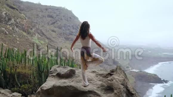 年轻女子在山上做瑜伽单腿站立俯瞰大海视频的预览图