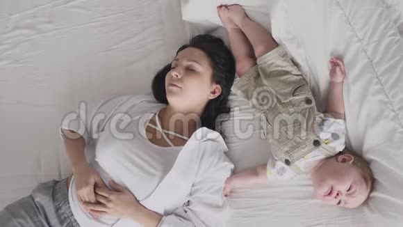 年轻疲惫的母亲睡在床上一个哭泣的小孩旁边视频的预览图