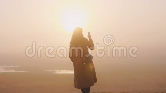 史诗般的剪影拍摄的快乐旅游女人拍摄智能手机视频惊人的粉红色雾日落景慢动作视频的预览图